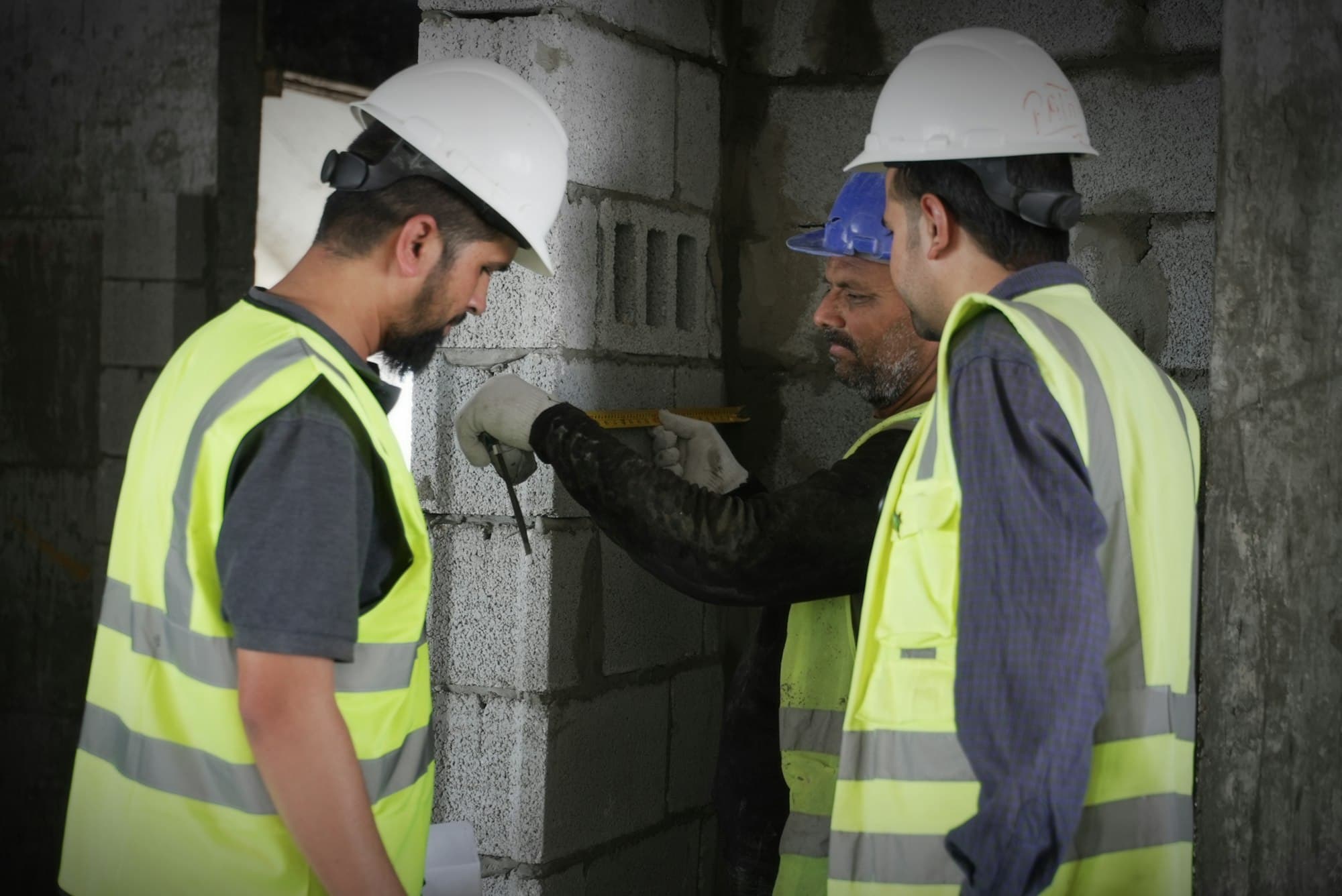 Three workers in hi-vis vests measuring and inspecting a block wall